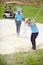 A chip off the old block. A young female golfer chipping her ball out of a bunker while her male partner looks on from