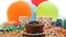 Chocolate birthday cake with a blue candle burning on rustic wooden table with background of colorful balloons, gifts