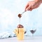 Chocolate brown ice cream in a regular yellow mug, on a blue background, the hand pours from a spoon topping. Unusual serve of