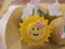 Chocolate cake, small, covered in yellow chocolate decorated with a smiling sun face served on a stick.