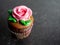 Chocolate cupcake with chocolate frosting decorated with a large pink rose and green leaves sitting on a black slate background