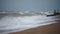 Choppy waves crash on sandy shore under overcast sky. Storm surge hits beach erosion barrier. Seagulls hover amidst