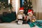 Christmas activity indoor: two boys in santa hats hold and considering boxes with gifts, sitting on a bed near a xmas tree with