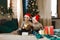 Christmas activity indoor: two boys in santa hats hold and considering boxes with gifts, sitting on a bed near a xmas tree with