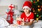 Christmas activity indoor: young child boy in santa hat hold gift and sitting on a bed with boxes with gifts near a xmas tree with