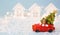 Christmas decor - red retro car on snow carries past white houses with lights garlands in bokeh Christmas tree with gift boxes on