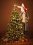 Christmas Family Decorate Xmas Tree. Child in Santa Hat putting Star on Top. Happy Father Together with Daughter indoors Portrait