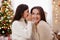 Christmas, family and motherhood concept - preteen daughter whispering secret to her happy mother in decorated living room with