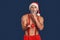 Christmas Freestyle. Young Santa Claus bare muscular upper body in hat standing isolated on blue holding proposal ring