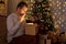 Christmas and magic concept - portrait of surprised young man opening gift box in decorated dark room with Christmas tree and led