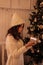 Christmas, New Year. Pretty young woman in white sweater and knitted hat  with  luminous gift box near christmas tree,  new year m