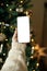 Christmas phone mock up. Hand holding smartphone with empty screen against stylish festive christmas tree with golden lights.