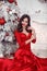 Christmas portrait of happy smiling girl in red dress sitting by snowy xmas tree, brunette with red lips, manicured nails and