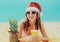 Christmas portrait of happy smiling young woman drinking fresh juice in red santa hat with pineapple lying on beach over blue