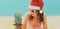 Christmas portrait of happy young smiling woman in red santa hat with funny pineapple lying on a beach together over sea