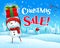 Christmas Sale! Cheerful snowman holds up gift present in Christmas snow scene winter landscape.