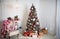 Christmas tree with red and white decor in a white living room with gifts in boxes, a chair with pillows and a blanket with winter