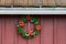 Christmas wreath made of artificial evergreen boughs and a light string, with red flowers and white snowflakes, on a red barn wall