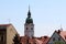 Church bell tower with analogue clocks on all sides and large metal cross on top rising high above city family houses rooftops