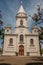 Church facade and belfry in front of a small cobblestone square with evergreen garden, in a sunny day at SÃ£o Manuel.