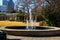 A circular water fountain in the garden surrounded by bare winter trees, lush green trees and plants, curved wooden benches