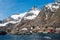 The city of Aa in Lofoten, Norway. Tiny red houses called rorbuer underneath steep snow covered mountain peaks and blue sky.