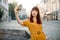 City portrait of cheerful young Caucasian girl, wearing yellow fashionable clothes, taking selfie on her smartphone