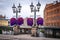 Cityscape view of purple flowers in flowerpot hanging from vintage lampposts. Industrial buildings in the background in Norrkoping