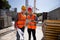 Civil architect and construction manager dressed in orange work vests and helmets discuss a building project on the