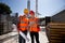 Civil architect and construction manager dressed in orange work vests and helmets discuss a building project on the