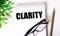 CLARITY is written in a white notebook next to a pencil, black-framed glasses and a green plant