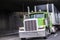 Classic big rig green semi truck transporting commercial cargo in covered black semi trailer running under the bridge across wide