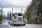 Classic big rig semi truck tractor transporting covered cargo on two flat bed semi trailer turning on the wet winding mountain