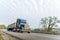 Classic blue big rig American bonnet semi truck driving with covered bulk semi trailer on the winding autumn road