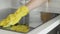 Cleaning cooktop cooking panel in kitchen with fat remover spray and yellow rag by a woman in yellow rubber gloves.