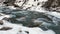 A clear mountain clear stream runs over the stones in winter. Amazing view of a stormy mountain river in the snow.