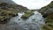 A clear, rocky stream flowing through a mountainous area with a cloudy sky.