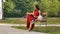 Clever Female Student in Eyesglasses and Red Long Dress Sitting on the Bench in the Park and Writing Something in a
