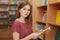 Clever young female with bobbed hairstyle, holds book in hands, poses in library or bookstore, dressed in casual t shirt, chooses