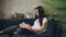 Clever young lady with long dark hair is reading book and holding cup of coffee sitting on couch in living room in