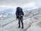 A climber with a backpack and trekking poles stands on a snowy ice trail