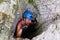 Climber girl fooling around on via ferrata, mimicking biting the wire cable, while exiting a cave through a very narrow opening