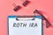 Clipboard with words roth ira near stationery items on pink desk