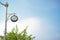 Clock hanging on a vintage pole, outdoors on a sunny day, blue clear sky background. Black dial showing the time outside.