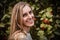 Close outdoor portrait of young blonde woman smiling to camera. She is posing next to a tree full of red apples.