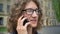 Close portrait of young handsome man in glasses with long curly hair talking on cell phone and smiling, standing on