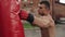 Close side view of muscular male boxer practicing punches and kicks with a punching bag being outdoors. Brown wall on