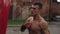 Close side view of muscular male boxer practicing punches and kicks with a punching bag being outdoors. Brown wall on