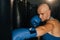 Close-up of anger bald boxer male training punches, beating punching bag in sport club with dark interior.