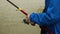 Close-up of an angler in a blue jacket spinning a reel handle on a red fishing rod against a river background. The